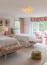 59 - Sissy Yellen, A vibrant girls' bedroom featuring two twin beds with scalloped white bedding, monogram details, and colorful chevron-patterned benches with acrylic legs. The room includes pink accents such as lamps, curtains, and armchairs, complemented by a crystal chandelier and large windows that fill the space with natural light. Rattan storage baskets are placed on a neutral carpet for added texture.