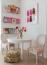 60 - Sissy Yellen, A charming kids' reading nook featuring a round white table and two cane-back chairs upholstered in colorful chevron fabric. Wall-mounted clear bookshelves showcase a selection of children's books, while a playful gummy bear art print hangs above. The space is accessorized with a vase of fresh pink flowers and woven rattan baskets for storage, creating a cozy and organized corner.